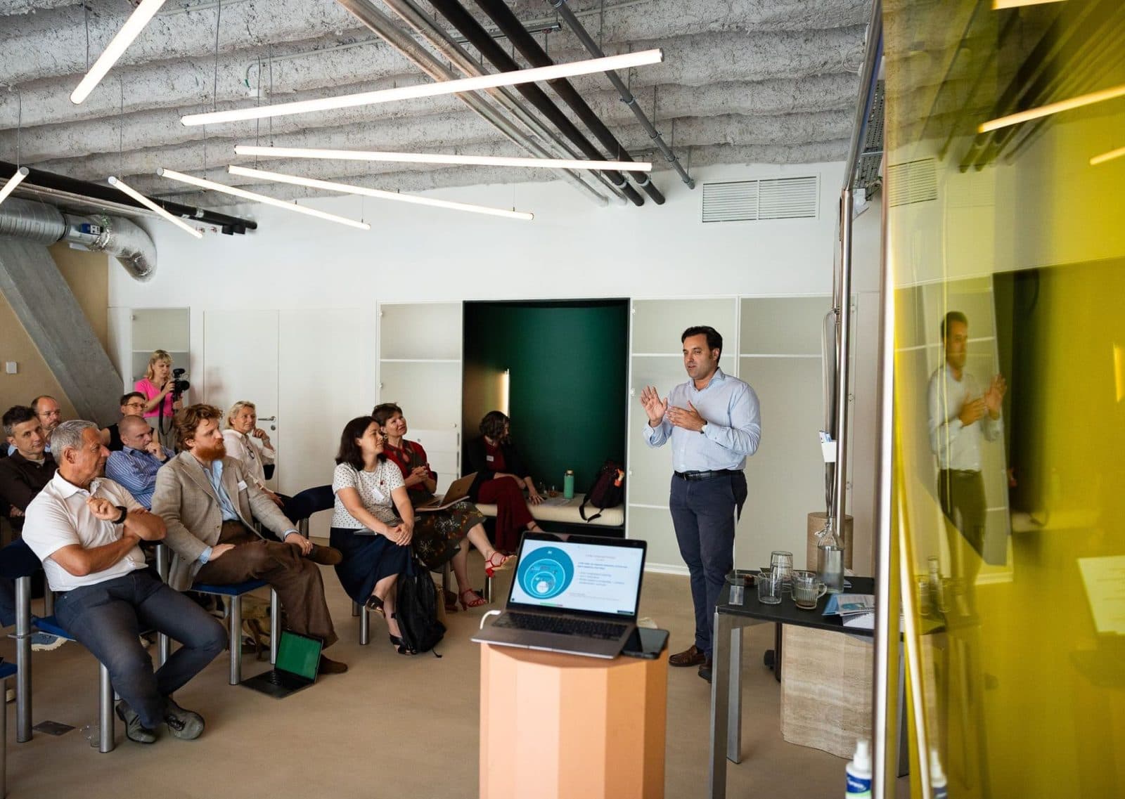 A trainer speaks to a room of seated participants during an AI workshop, with a slide on large language models open on a laptop in the foreground
